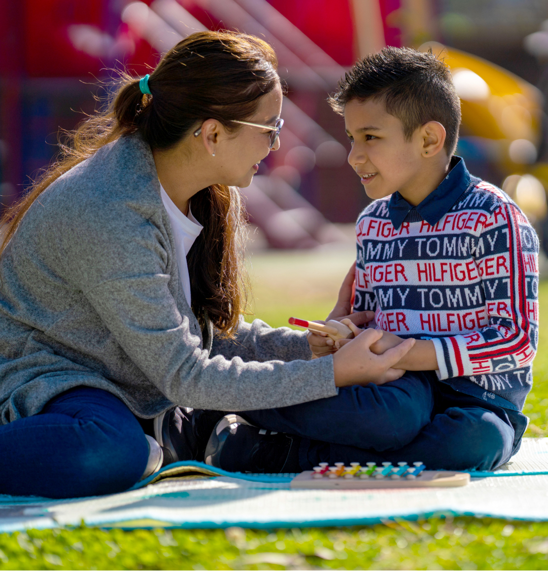 A mother is sitting on a blanket with her primary-school aged son. They are looking at each other smiling and the mother is holding the boy’s hands. We can see a toy xylophone in front of the boy. It is a sunny day.