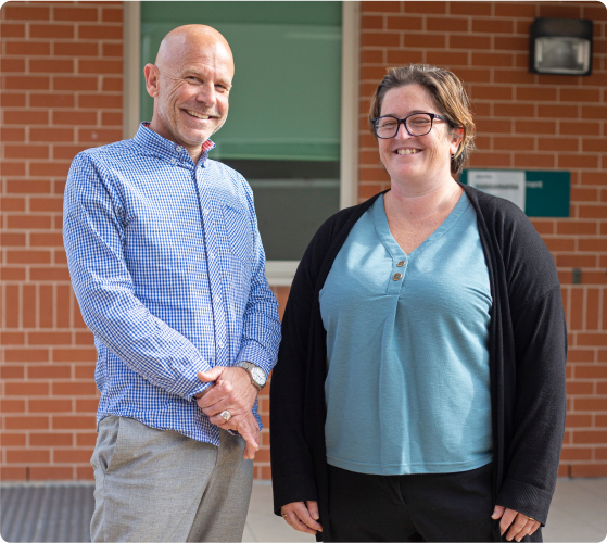 A man and woman are standing side by side in front of a brick building. They are looking at the camera smiling. The man is Aaron. He is wearing a check collared shirt. The woman is wearing a black cardigan and green shirt.