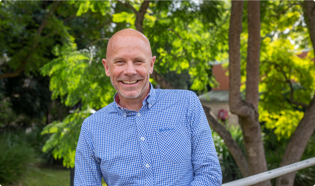 A man in a blue checked shirt is in a garden, looking at the camera, smiling.