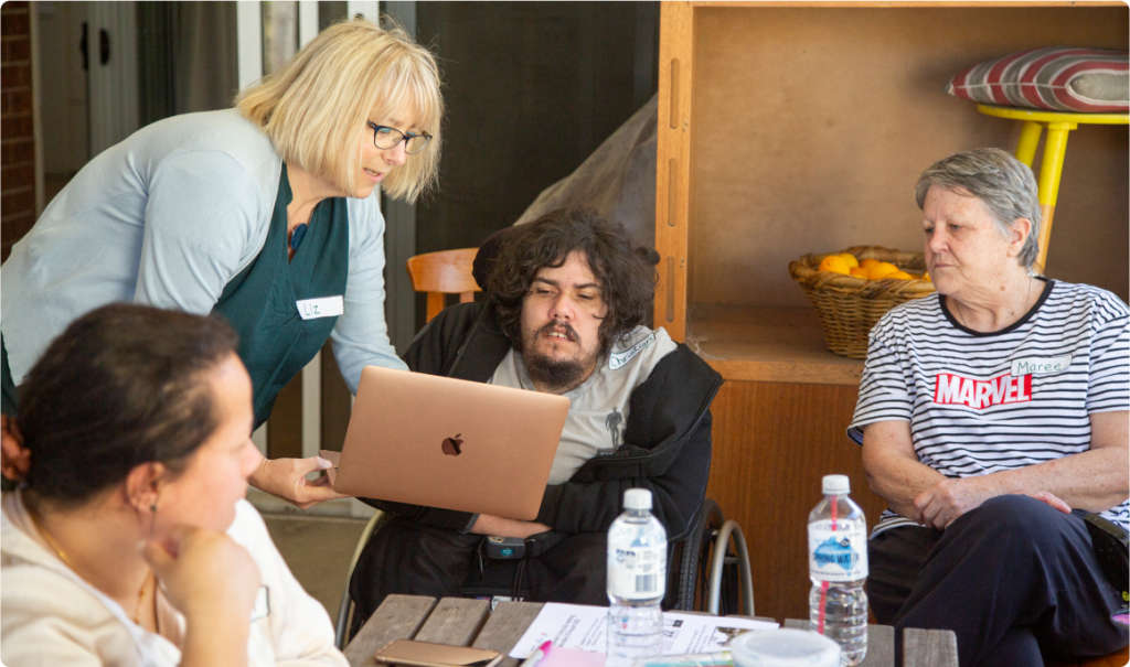 A woman is holding a pink Apple Mac Air laptop and showing it to a man in a wheelchair. Two other women either side are looking on.