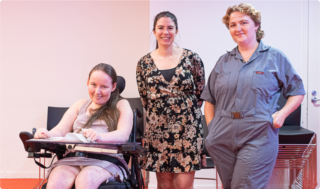 A woman is sitting in a motorised wheelchair with a table attached. Two other women stand next to her. Everyone is smiling.
