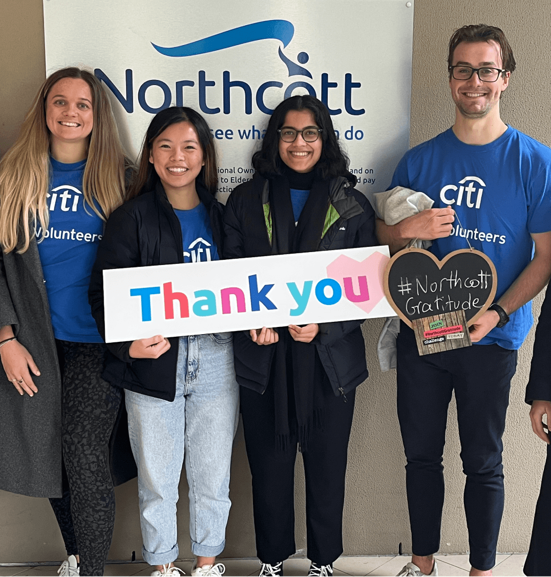 Four people standing in front of a Northcott sign. They are all wearing blue Citi volunteers t-shirts and holding a colourful thank you sign. One person is holding a heart shaped blackboard that says "hashtag Northcott Gratitude"
