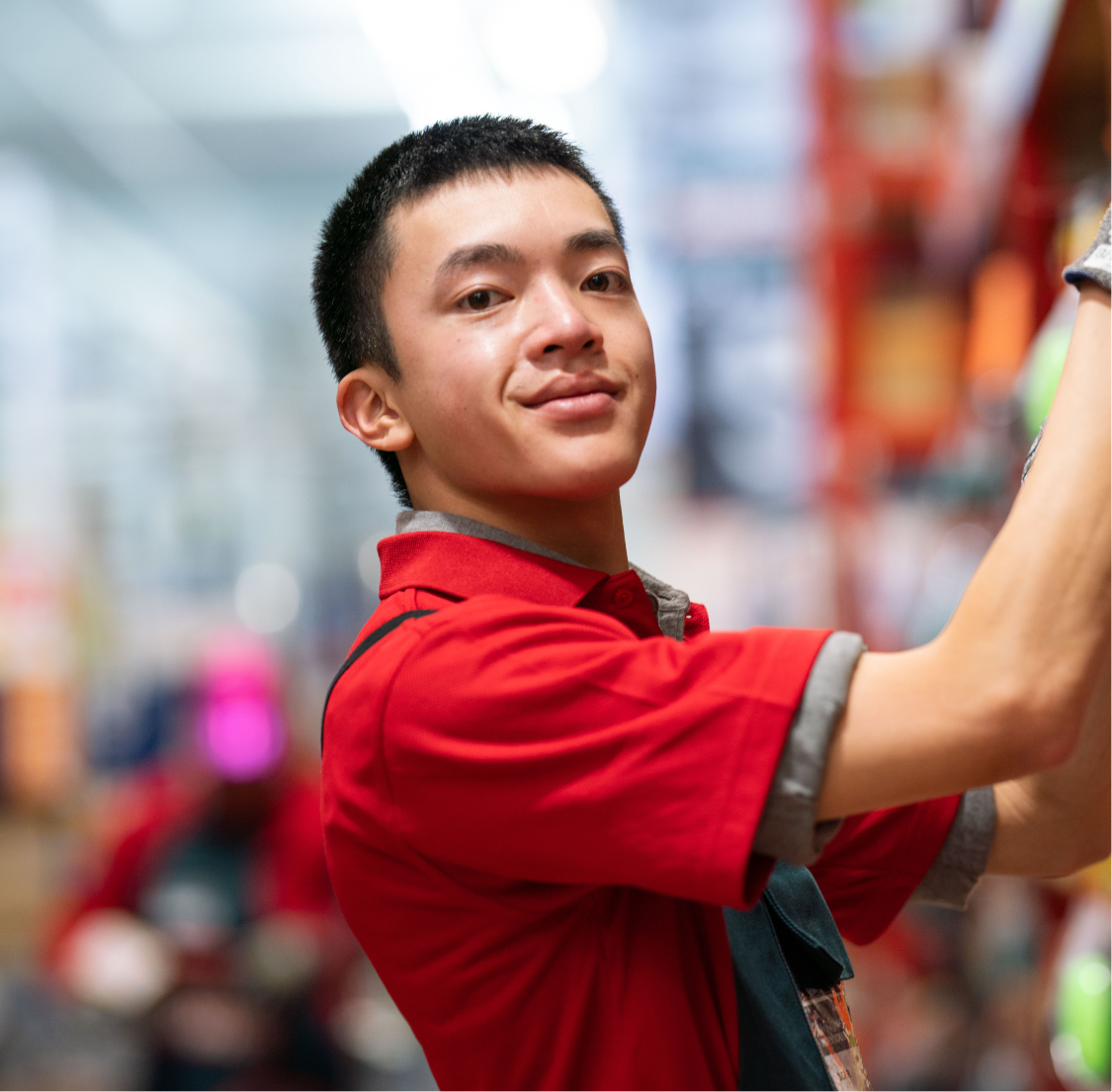 A young man, Oscar, is standing facing a shelf, with his head turned towards the camera. He is wearing a red shirt and is smiling slightly at the camera. It looks like he is working in an aisle.