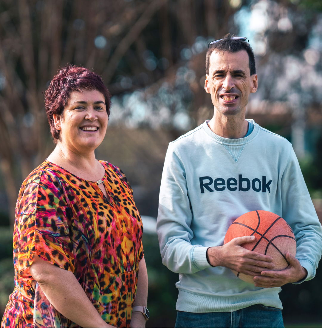 A woman in a colour red and orange top is standing next to a man in a Reebok jumper holding a basketball. Both people are looking towards the camera.