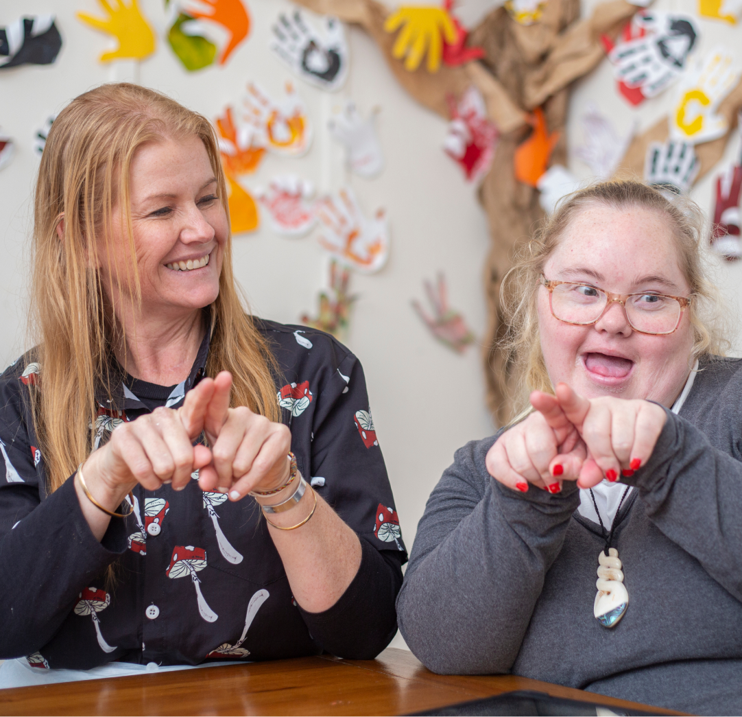 A woman with orange hair, Rachel, wearing a black shirt with red and white mushrooms on it is sitting next to and looking at a younger woman with Down Syndrome wearing glasses. Both women are doing Key Word Sign and smiling. On the wall behind the women is a wall with a picture of tree on it and colourful hands prints stuck to it.