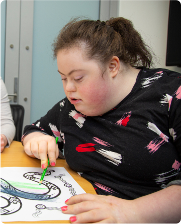 A young woman is seating at a table with some colouring-in pictures on it. She is holding a green pencil and colouring-in what looks like a snake.