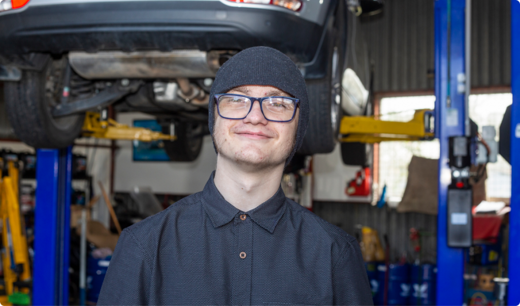 A portrait of a young man, Sean, smiling at the camera. He is in front of a car hoisted off the ground. He is wearing dark bean, collared shirt buttoned to the top and square glasses.