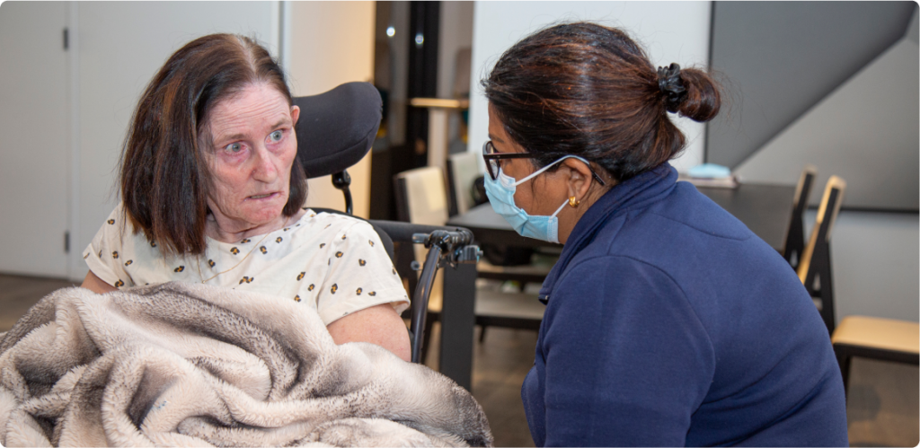 A woman is in a wheelchair and covered with a blanket. Another woman in a facemask and wearing a blue jumper is sitting next to her, wearing a face mask. Both women are looking at each other. You can see a dining table and cupboard in the background.