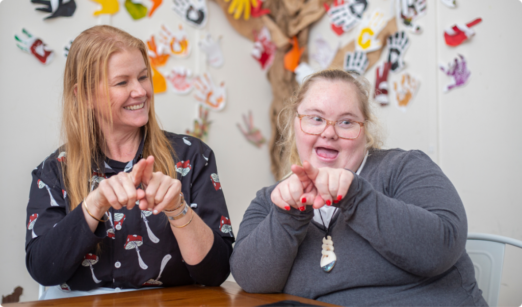 A woman with orange hair, Rachel, wearing a black shirt with red and white mushrooms on it is sitting next to and looking at a younger woman with Down Syndrome wearing glasses. Both women are doing Key Word Sign and smiling. On the wall behind the women is a wall with a picture of tree on it and colourful hands prints stuck to it.