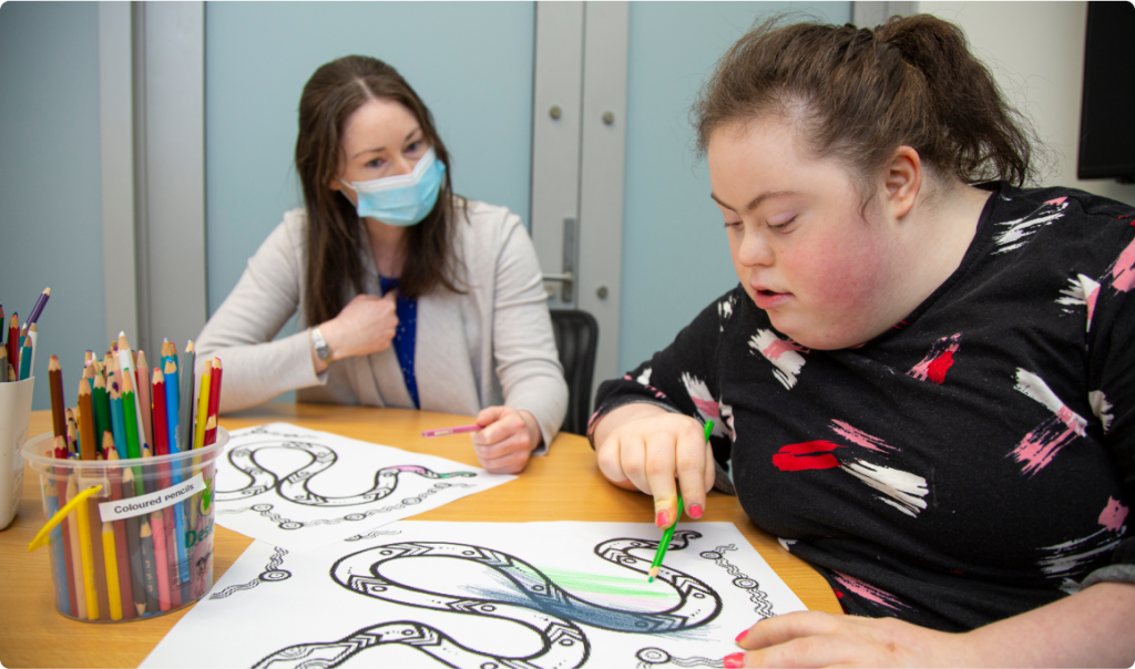 Two women are seating at a table with some colouring-in pictures on it with a tub of coloured pencils. One woman is wearing a face mask and holding a pink pencil. The other woman is holding a green pencil and looking down at the drawing of a snake.