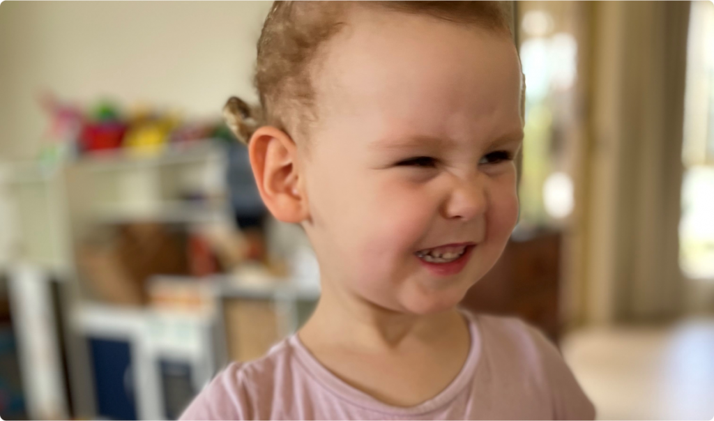 A young girl, Harper, dressed in pink and smiling with a scrunched up nose. She is wearing a pink t-shirt.