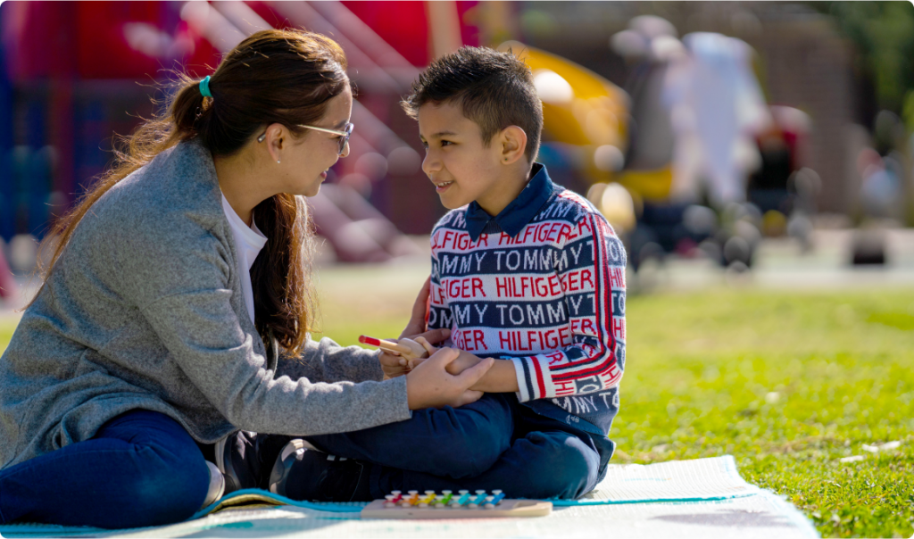 A mother is sitting on a blanket with her primary-school aged son. They are looking at each other smiling and the mother is holding the boy’s hands. We can see a toy xylophone in front of the boy. It is a sunny day.