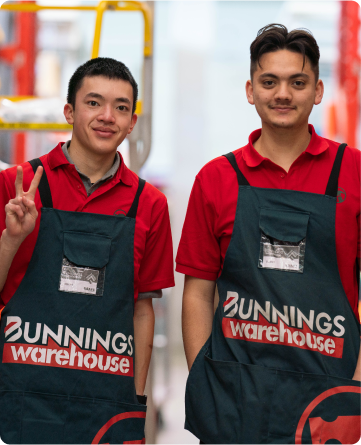 Two young men, both wearing red polo shirts and green Bunnings Warehouse aprons, are standing together in an aisle. They are looking at the camera smiling. On guy is giving the peace symbol with his fingers.