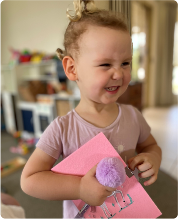 A young girl, Harper, dressed in pink and smiling with a scrunched up nose. She is holding a pink card.
