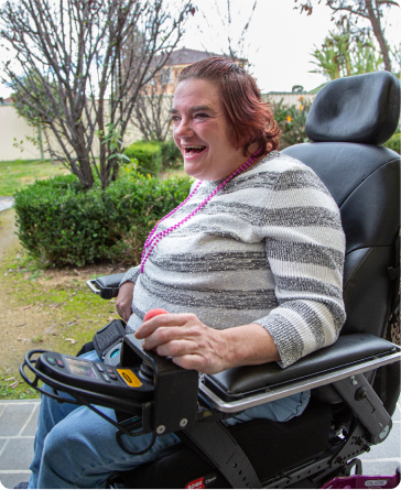 A woman is in a motorised wheelchair, holding a red joystick controller. She is smiling widely and looks happy. She is in a garden.