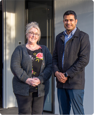 A woman and a man are standing outside a home, smiling at the camera. The woman is wearing a black cardigan, black top with colourful flowers on it and black pants. She has blond hair and wears glasses. The man is from the sub-continent and is wearing a black coat, blue checked shirt and blue pants.