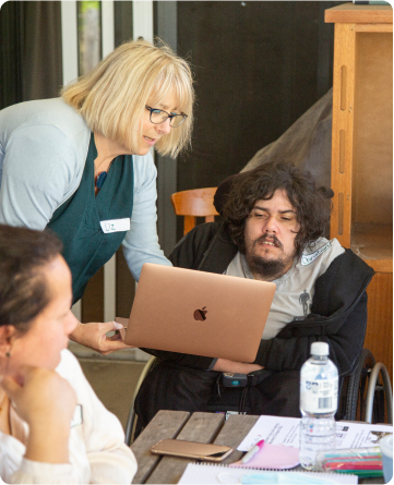 A woman is holding a pink Apple Mac Air laptop and showing it to a man in a wheelchair. Two other women either side are looking on.