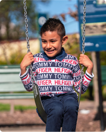 A young primary-school aged boy is sitting on a swing. He is wearing a striped blue and white Tommy Hilfiger jumper with red and white writing. He is smiling.