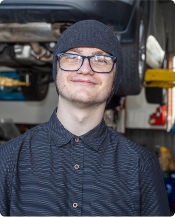 A portrait of a young man, Sean, smiling at the camera. He is in front of a car hoisted off the ground. He is wearing dark bean, collared shirt buttoned to the top and square glasses.