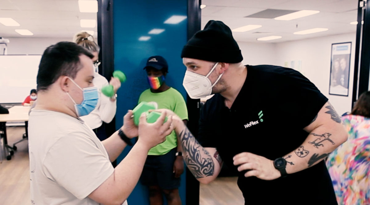 Two men both wearing face masks and looking at each. One man is passing a hand weight to the other man who is hoping it with two hands.