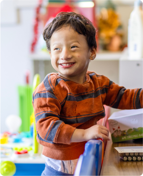 A two-year old child is standing and smiling. He is wearing a red and blue striped top. The background is blurred out by colourful.