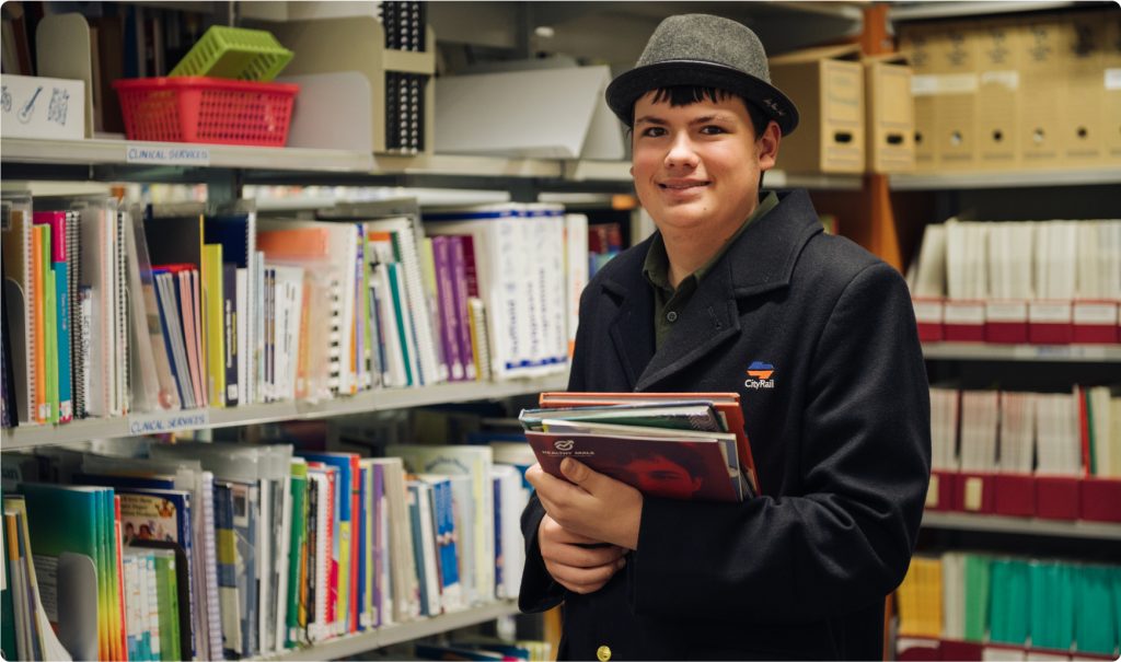 A young man in a large woollen coat and hat is holding a pile of books and standing in front of book shelves. It looks like a library. He is smiling at the camera.