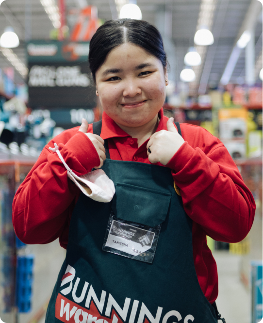 A young woman of Asian heritage is in a Bunnings Warehouse uniform. She is standing in a tools aisle and is giving the camera thumbs up. She is smiling.