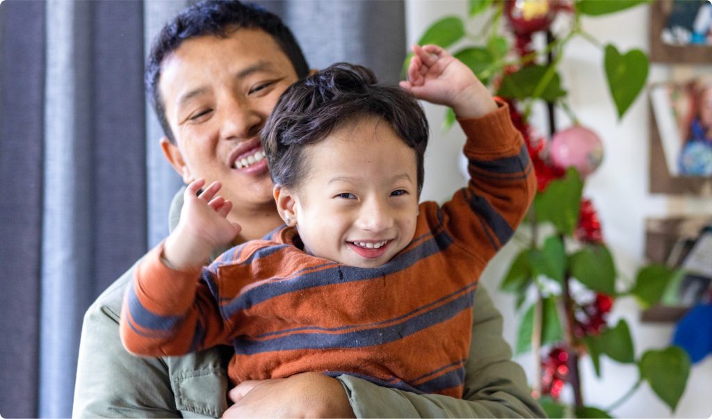 A young boy is being hugged by a man. The young boy has his hands up in the air and is smiling. The man is smiling.