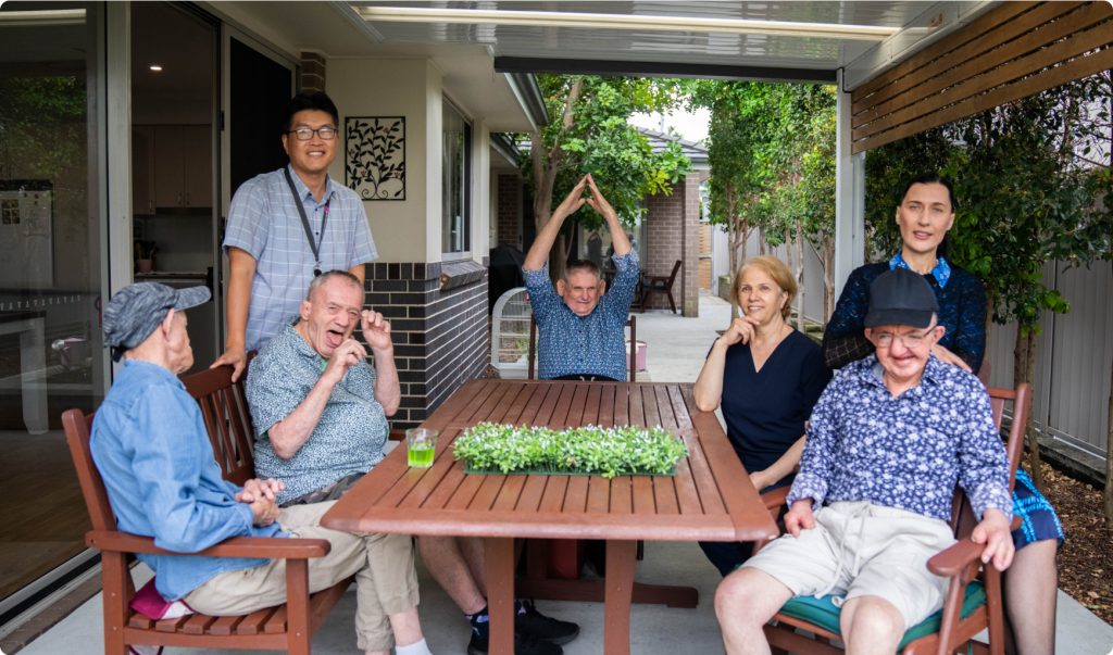 A group of people, four older men, two women and a young man and seated and standing around a wooden outdoor table in the outdoor area of a house. Everyone looks happy. Not everyone is looking at the camera.