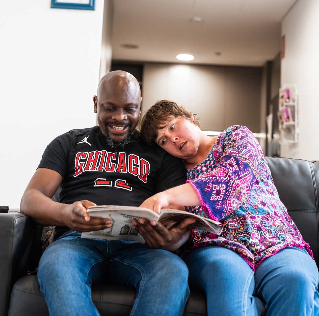 A man and a woman are seating on a couch looking at a magazine. The woman is resting her head on the man's shoulder, while pointing at something in the magazine. The man is smiling.