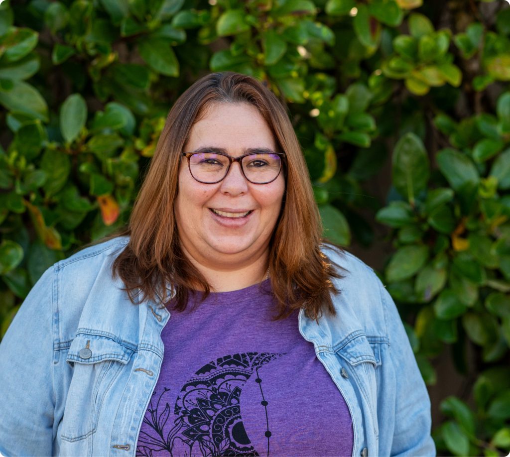 A headshot of a woman wearing a denim overshirt and a purple t-shirt. She is smiling.