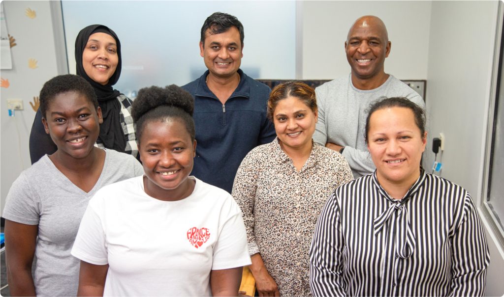 A group shot of seven people, all looking at the camera, smiling. They are a culturally diverse group.