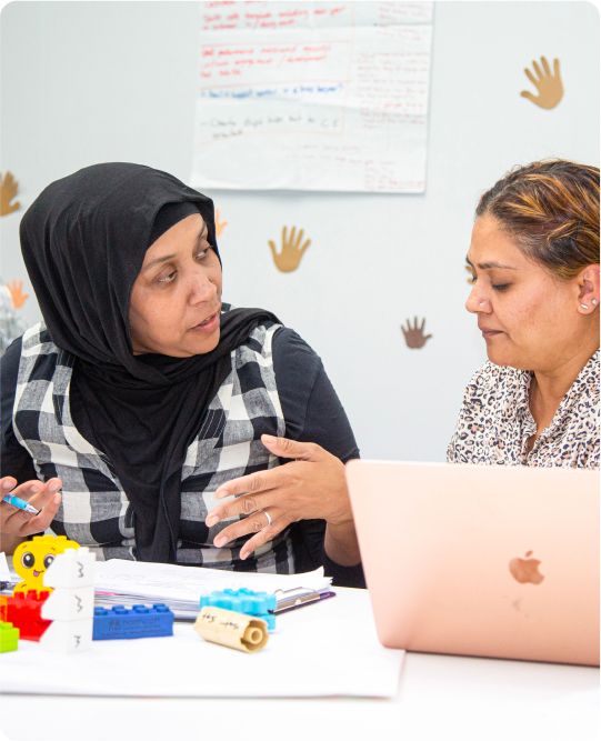 Two women an intently discussing something, while seated a desk with a laptop, notepad and lego resources in front of them. They are from culturally diverse backgrounds.