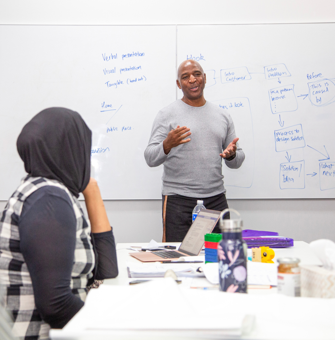 A man is standing in front of a whiteboard, explaining something. A woman is seated a desk is looking towards him. We see her back. Both are from a cultural diverse background.