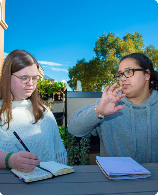 Two young women are seated at a table outside a building. They are talking and have notebooks in front of them.