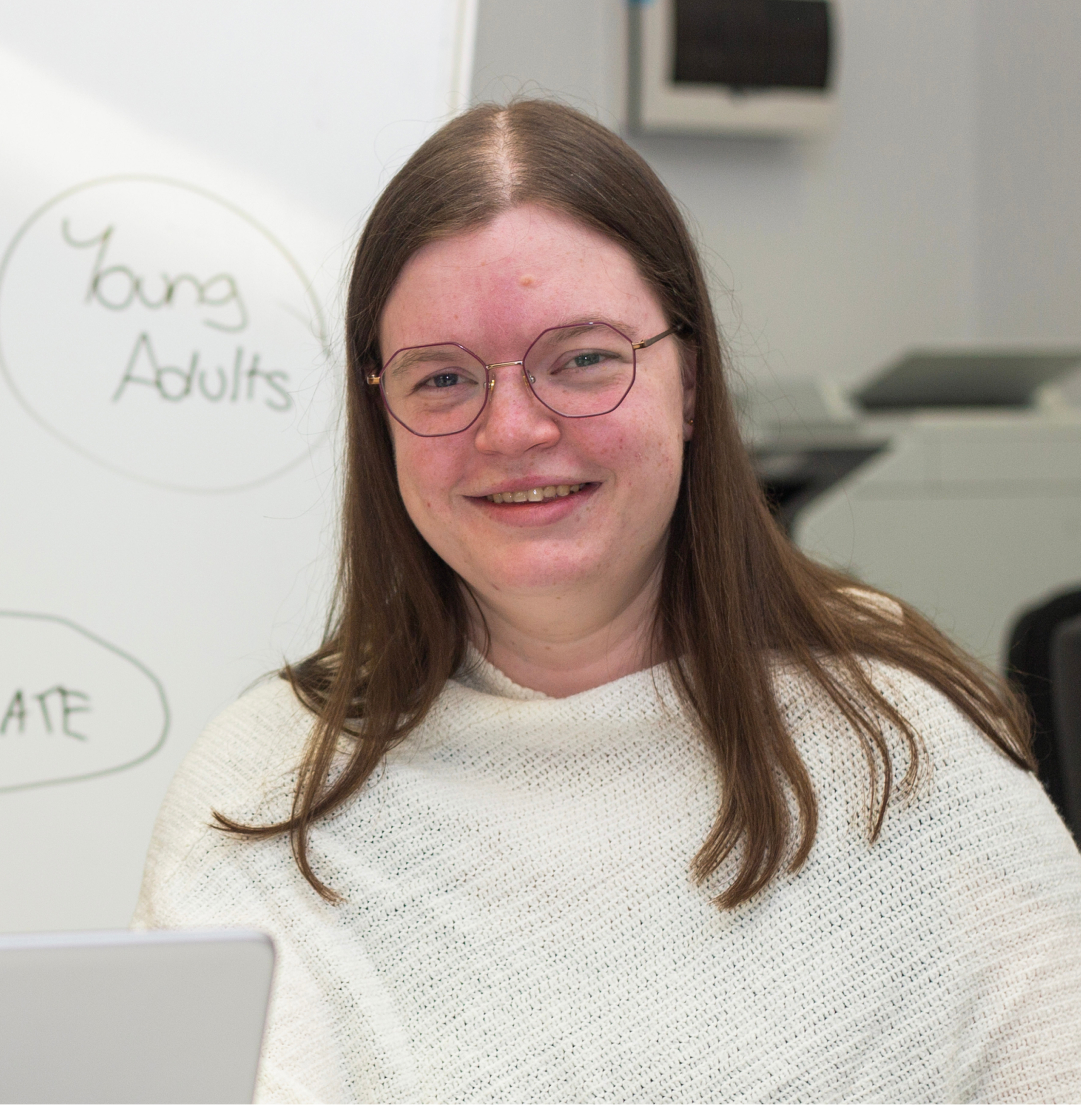 A young woman is smiling at the camera. She is wearing glasses and a white top. She is in front of a white board.