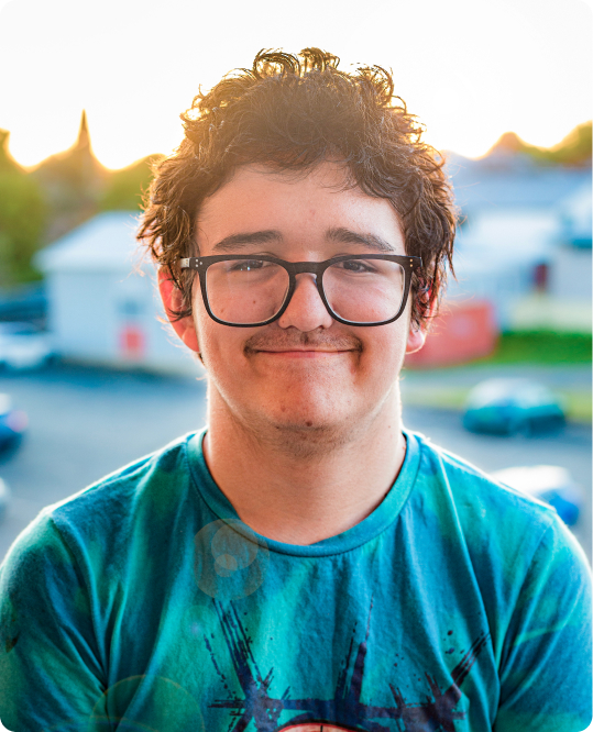 A young boy with glasses is looking straight at the camera. The sun is setting behind him. He is wearing a blue t-shirt and glasses.