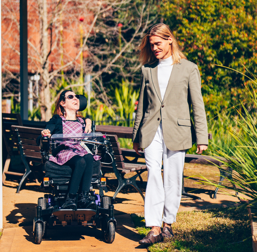 A woman is in a motorised wheelchair. A man with long blond hair and a moustache is walking beside her. They are outside in a park. They are chatting and smiling at each other.