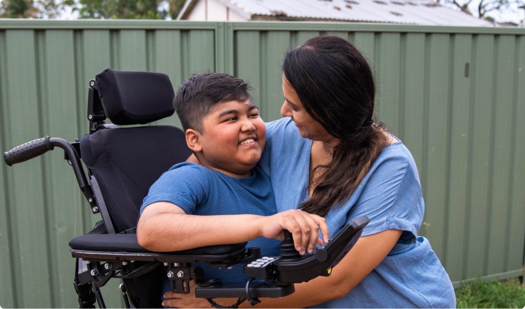 A teenage boy is in a standing wheelchair being hugged by a woman. They are smiling at each other.