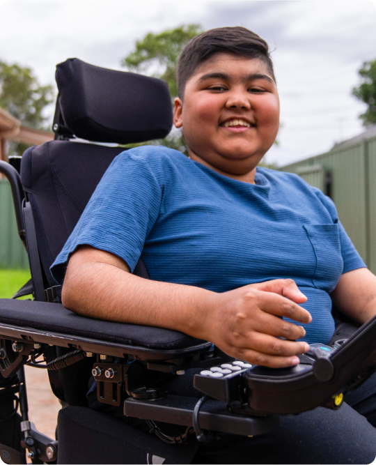 A teeange boy is in a standing wheelchair. His hand is on the controller. He is smiling at the camera.