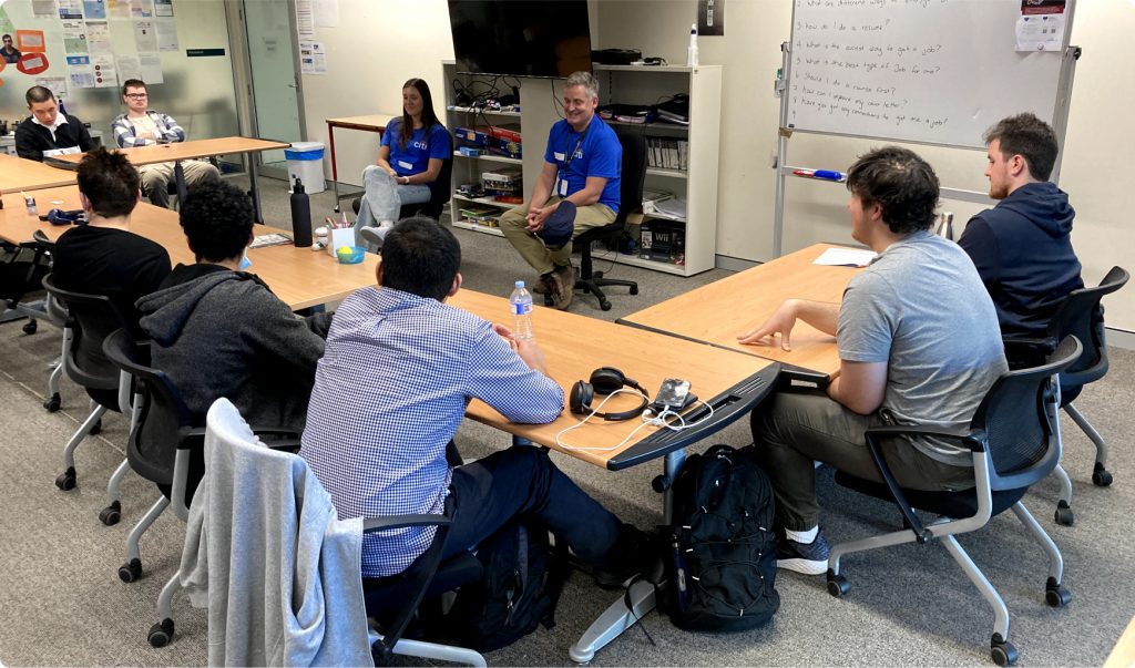 Two people in blue Citi T-shirts are seated in front of a room, talking with a grou pof young men who are all seated at desks in a U-shape.