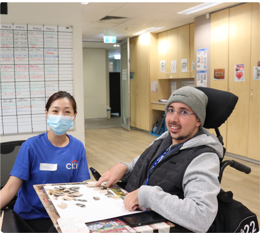 A man in a wheelchair is working on a collage, while a woman wearing a face mask and a t-shirt that says Citi is looking on. They are both looking at the camera and look happy.