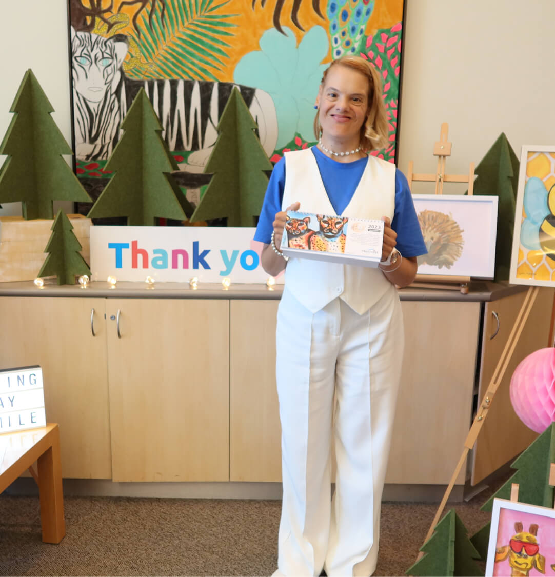 A young woman is dressed in white slacks and a white vest with a blue t-shirt. She is hold a desk calendar with a drawing of tigers on it. She is standing in front of artworks and a sign that says 'Thank you'